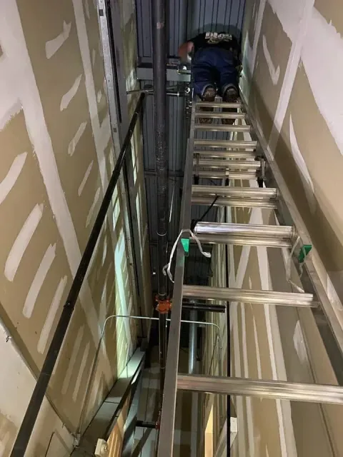 Worker on a ladder in a narrow unfinished hallway near exposed wiring and wall panels