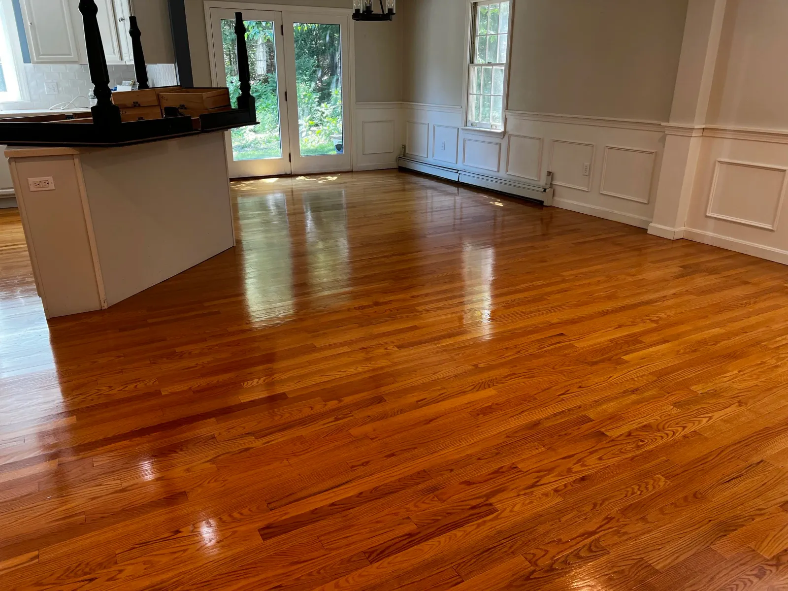 Empty room with polished wooden floors, white walls with decorative paneling, large windows, and a glass door leading outside.