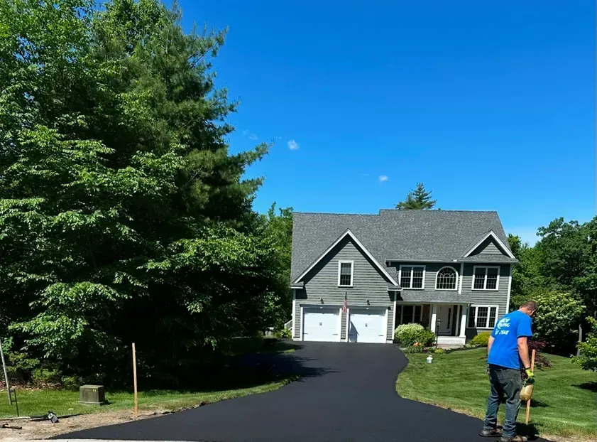A worker in a blue shirt standing near a newly paved asphalt driveway in front of a gray two-story house.