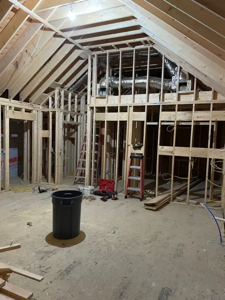 A wide-angle view of a room under construction with exposed wooden wall studs, ceiling rafters, and flexible HVAC ducts.