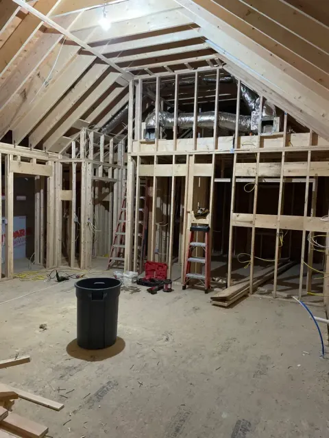 A wide-angle view of a room under construction with exposed wooden wall studs, ceiling rafters, and flexible HVAC ducts.