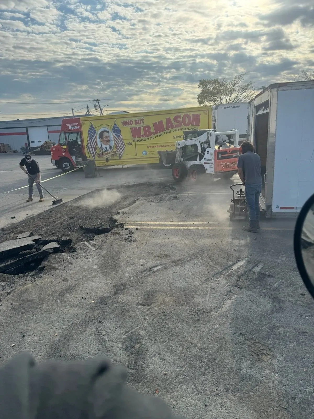 Damaged road with debris, workers, and a yellow truck stopped near a roadside building at sunset.