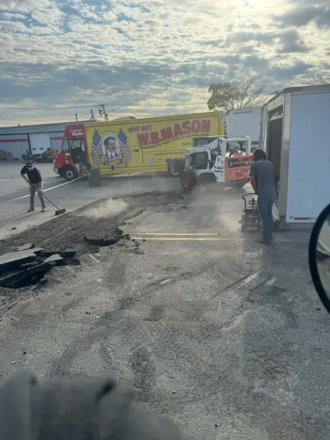 Damaged road with debris, workers, and a yellow truck stopped near a roadside building at sunset.