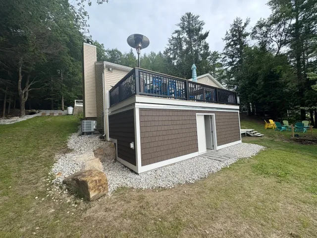 A two-story house with a deck on top, decorated with outdoor furniture and an umbrella, situated in a wooded outdoor area with Adirondack chairs in the background.