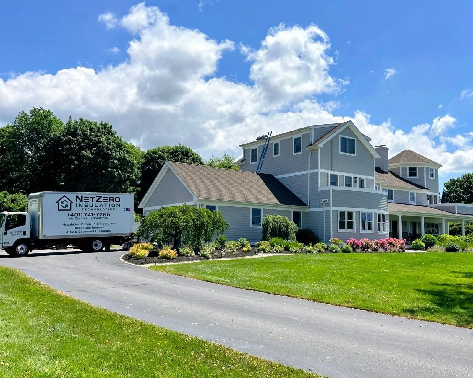 A residential neighborhood with a white house, a yard with flowers, and a sign for insulation services in front of the house. There are trees, a blue sky with clouds, and a paved driveway.