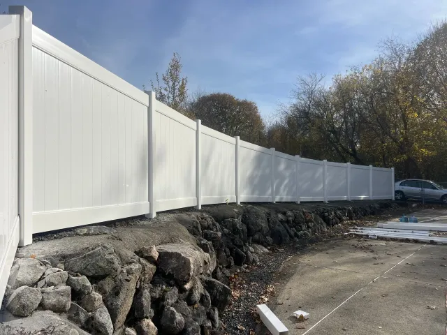 White vinyl fence along a rocky ledge, trees in the background, a sunny day.
