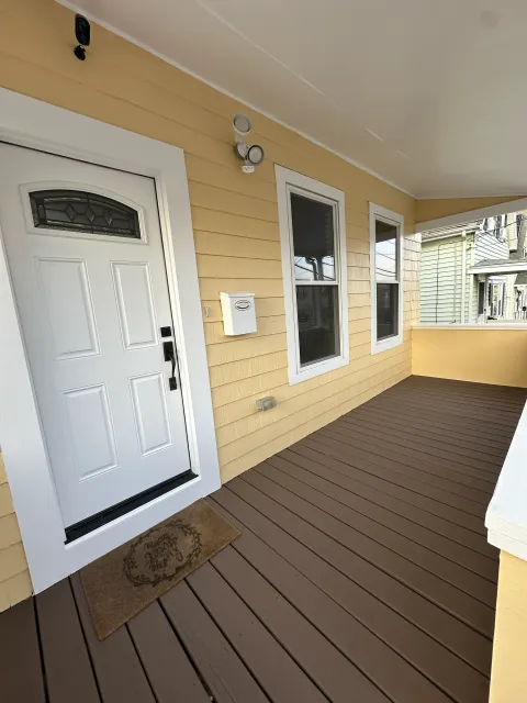 Front porch of a house with yellow siding, white door, three windows, brown deck flooring, and a doormat in front of the door.