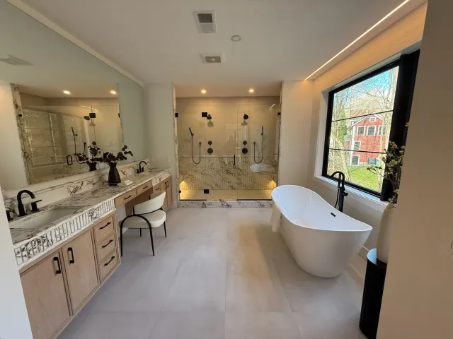 Modern bathroom with double sink vanity, large mirror, freestanding bathtub, and walk-in shower with glass enclosure and multiple shower heads, illuminated by natural light from large window.