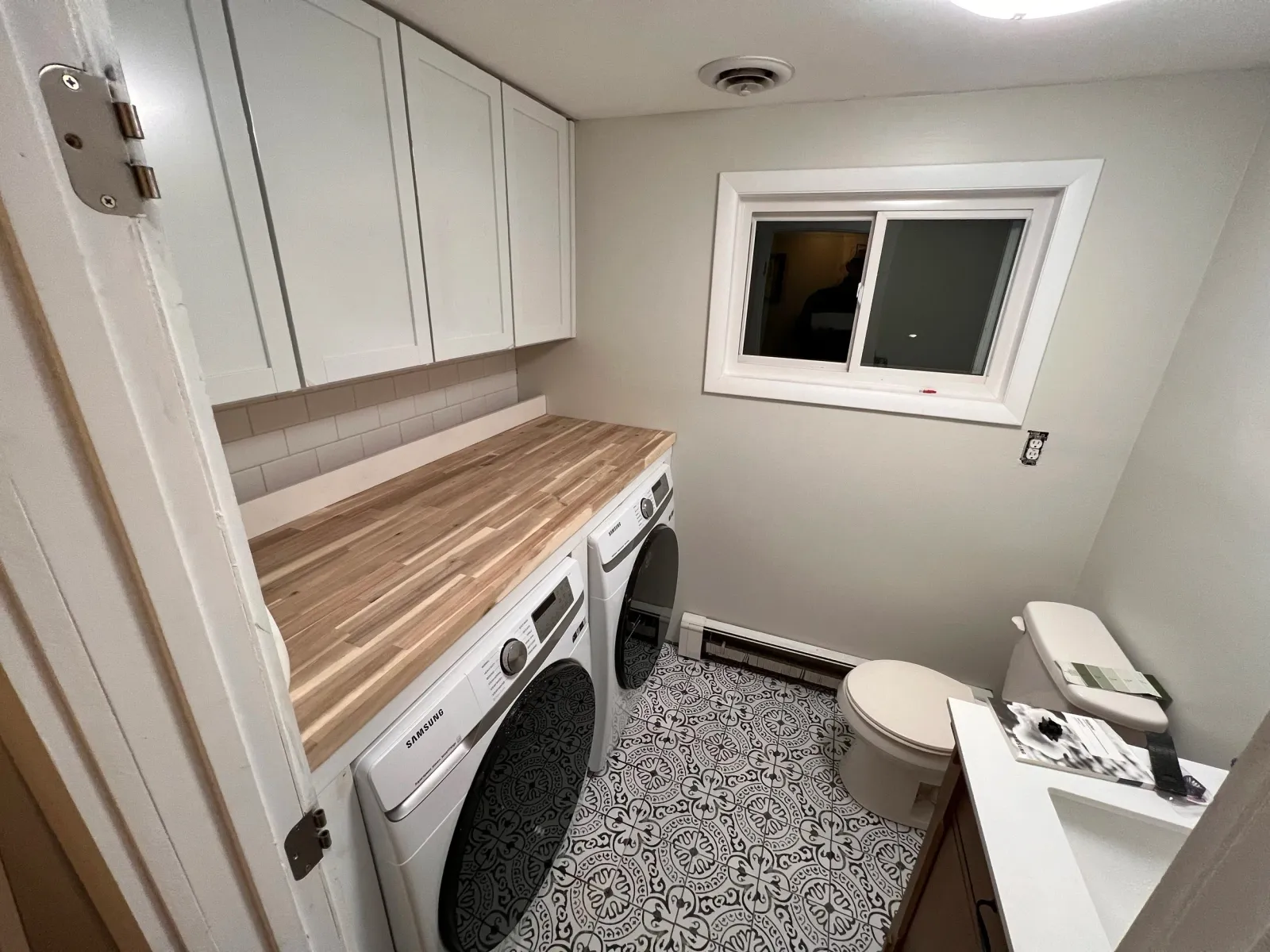 Small laundry room with a washer and dryer, a wooden countertop, white cabinets, a window, a toilet, and a patterned floor.