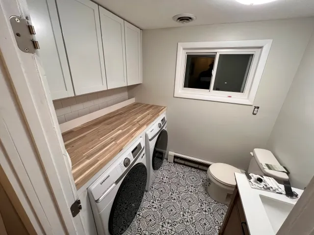 Small laundry room with a washer and dryer, a wooden countertop, white cabinets, a window, a toilet, and a patterned floor.