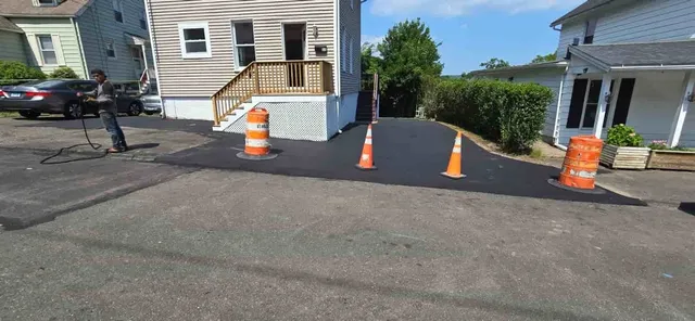 Orange traffic cones block a driveway beside a white house and street, with a dark car on the left.