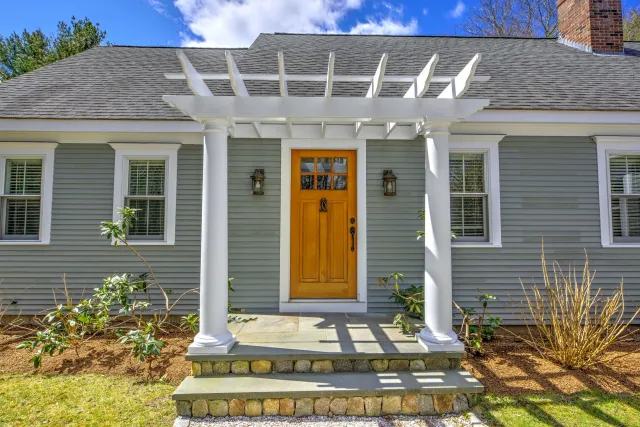 Front view of a house entrance with a wooden door framed by white columns supporting a white pergola. There are two windows on either side of the door, garden plants, a small set of stairs, and a brick chimney on the roof under a partly cloudy sky.