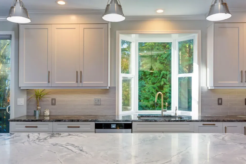 Modern kitchen with white cabinets, marble island, pendant lights, and a bay window above the sink.