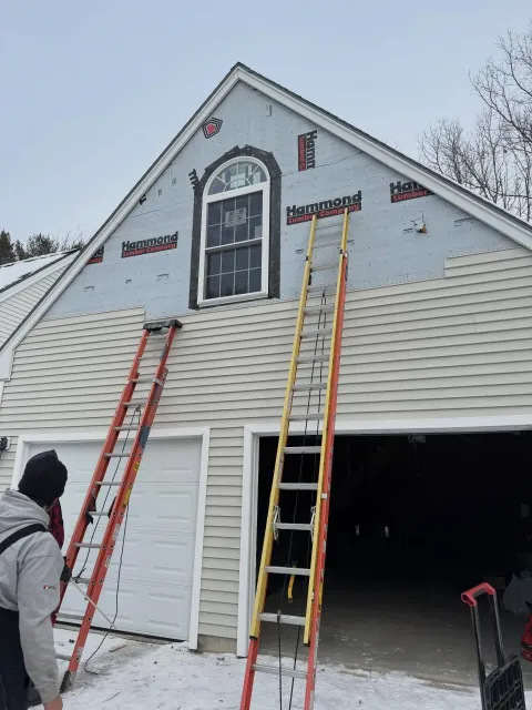 Two ladders against a house under repair, with workers below and open garage on the right.