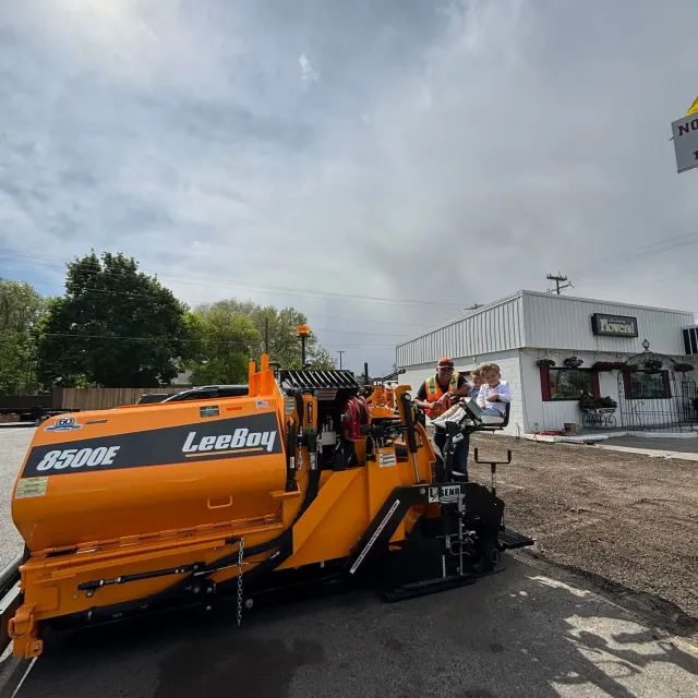 Orange road paving machine in front of a white building, under cloudy skies. Crew members visible.