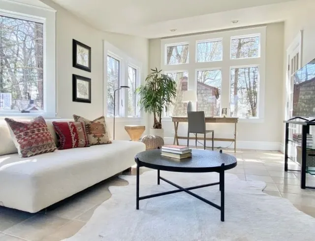 Bright living room with large windows, white walls, a white sofa with patterned pillows, a black round coffee table with books, a wooden chair, a desk with a chair, and indoor plants.