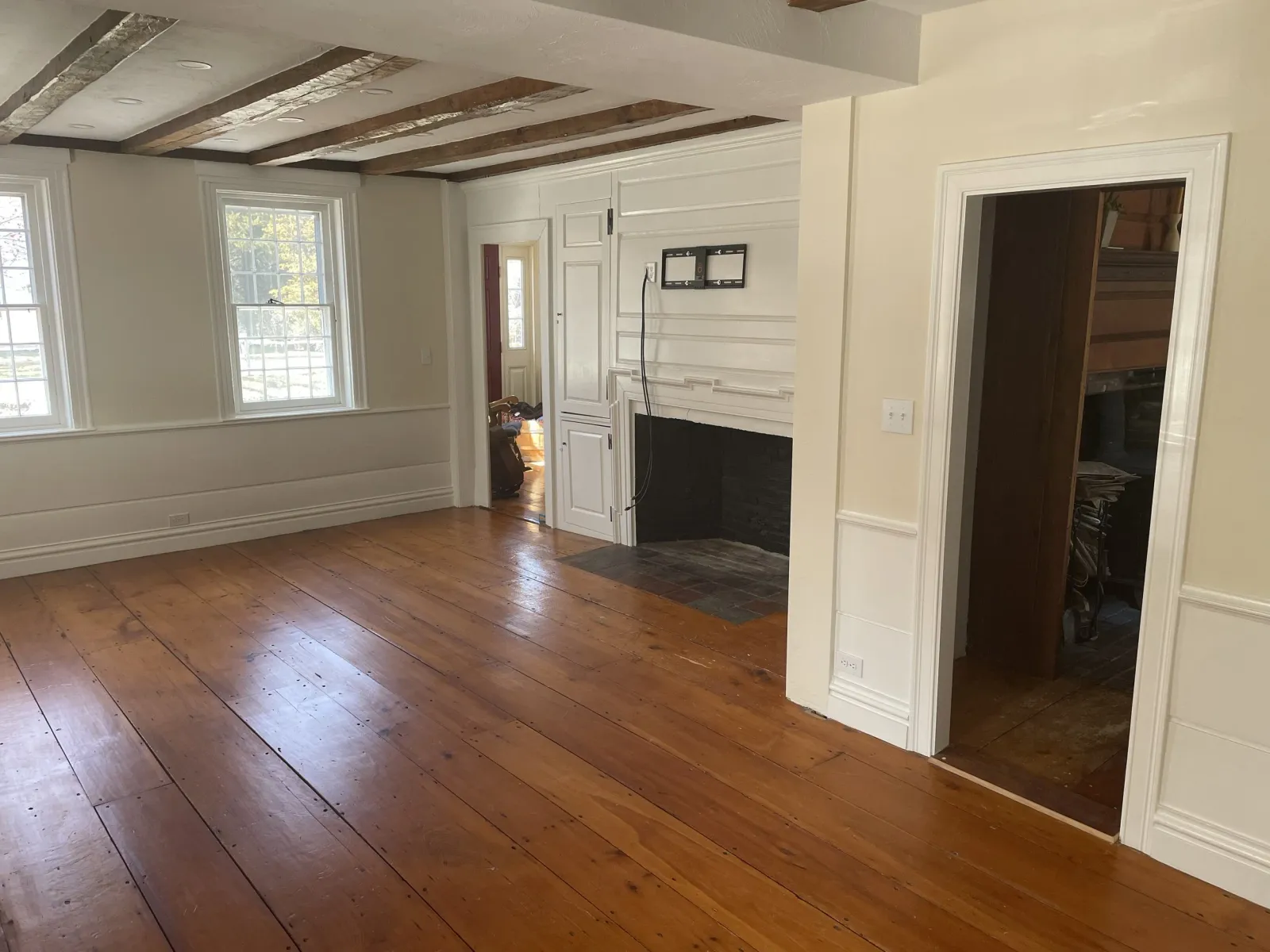Empty living room with hardwood floors, white walls, two large windows, a white painted fireplace, and an open doorway leading to a storage area.