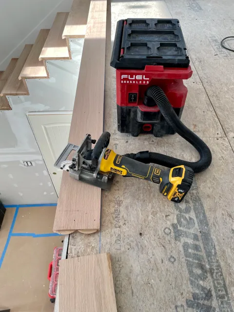 A woodworking workspace with a cordless circular saw, a red shop vacuum, and wooden planks on a workbench.