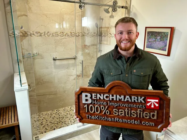 A wooden Benchmark “100% Satisfied” sign in a finished bathroom with a marble walk-in shower