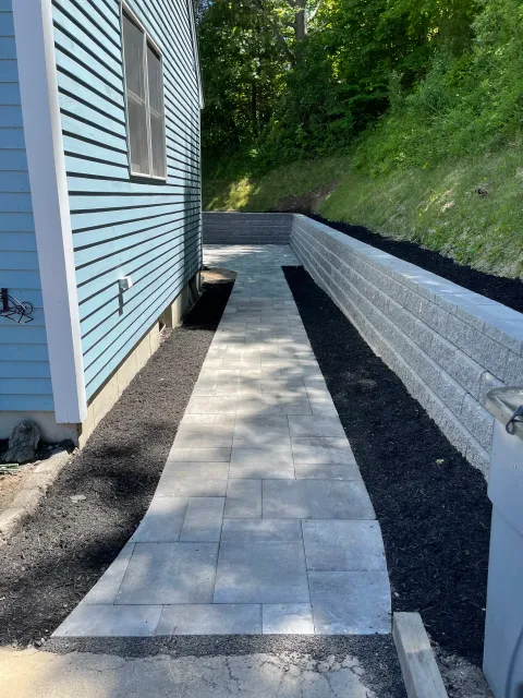 A newly layed Techobloc paver walkway leading to the side of a house with light blue siding and a window, flanked by a long low retaining wall made of gray bricks and a flower bed with black mulch along a grassy slope.