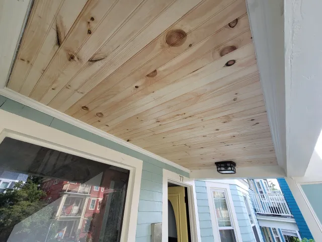 Porch ceiling with newly installed wood planks, light blue house exterior, window, and partial view of neighboring houses on Montebello Street, Jamaica Plain.