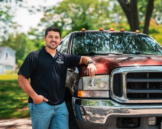 Man leaning on a red pickup truck in a tree-lined driveway, smiling at the camera