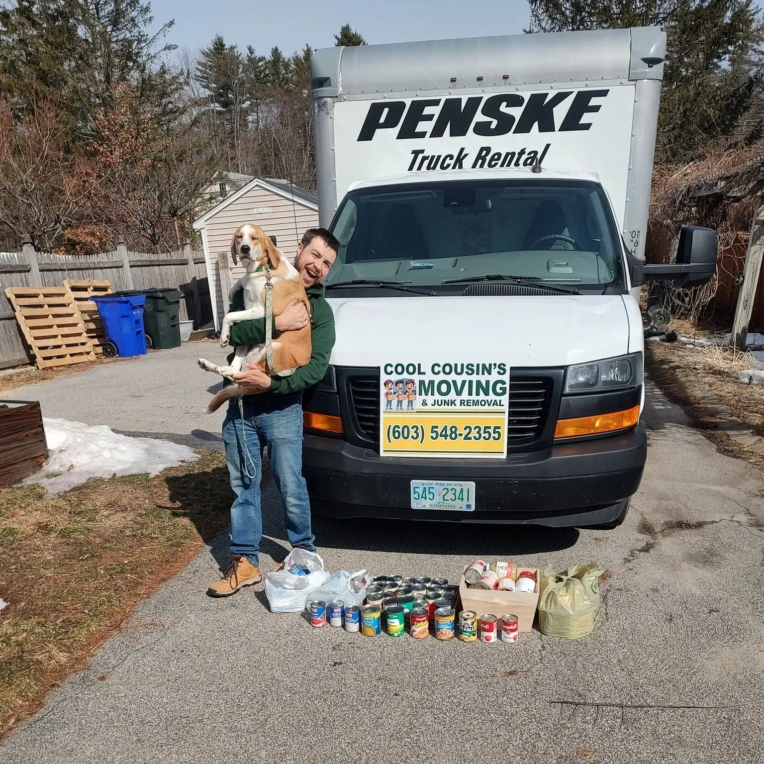 Mover holding a dog in front of a Penske truck, with canned goods lined up for a donation drive.