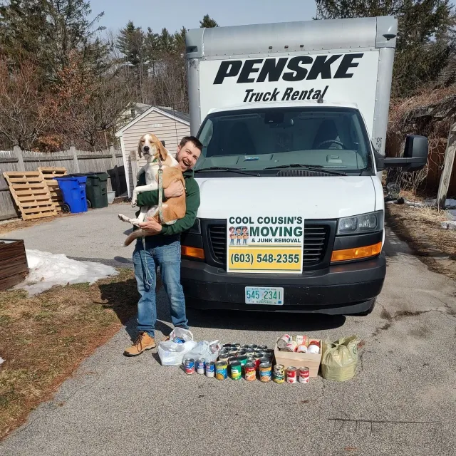 Mover holding a dog in front of a Penske truck, with canned goods lined up for a donation drive.