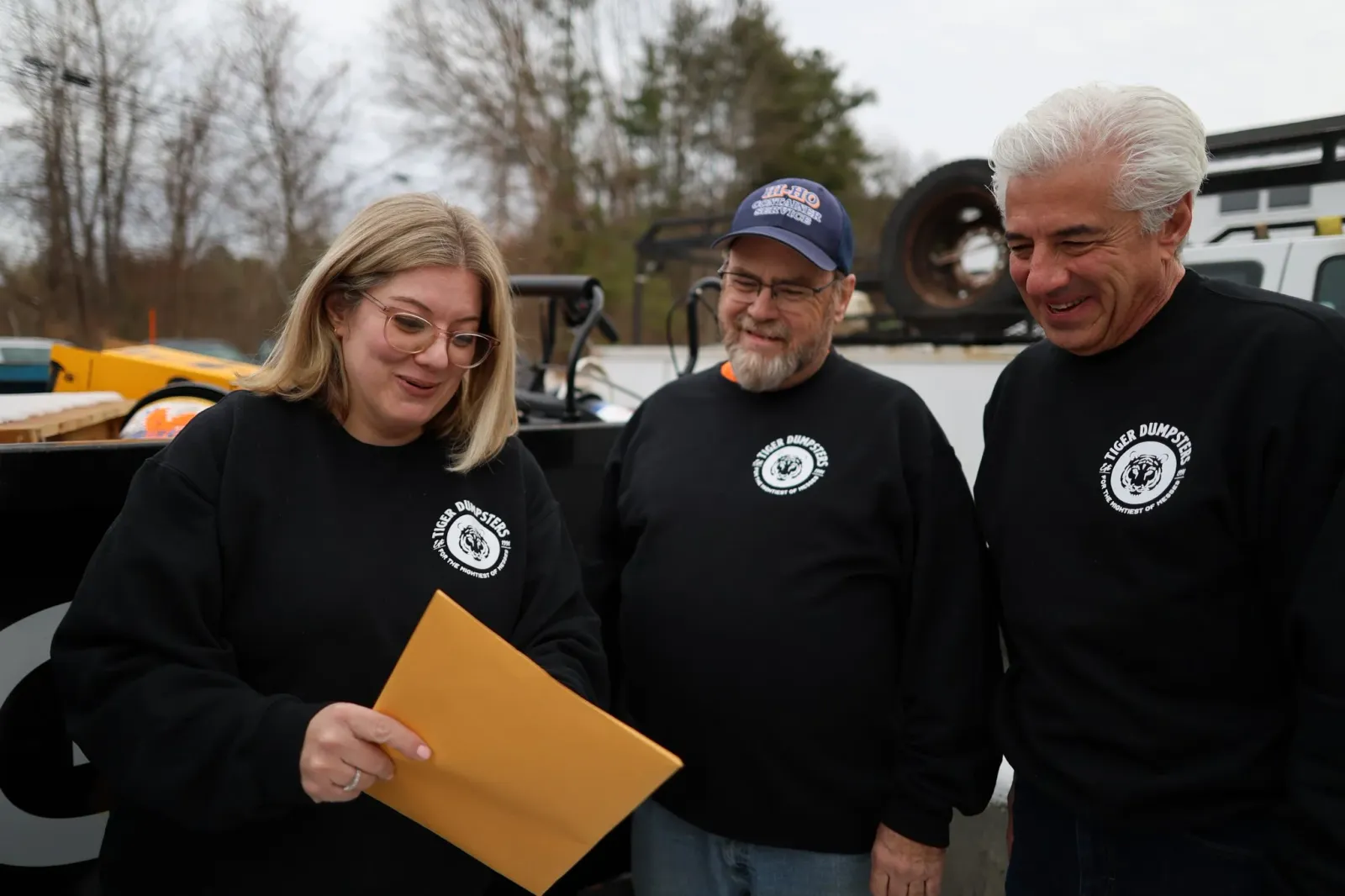 Three people standing outdoors, wearing black sweatshirts with a logo that reads 'Tiger Dumpster.' They are looking at a piece of paper or folder that the woman on the left is holding, smiling.