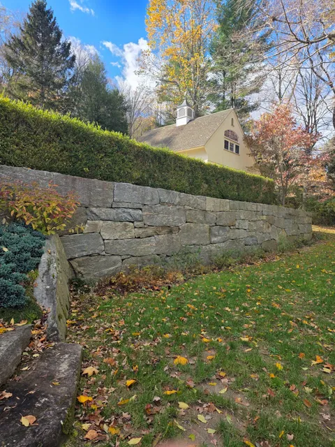 Stone wall topped with green hedge in front of a light yellow building, autumn foliage scattered on the grass.