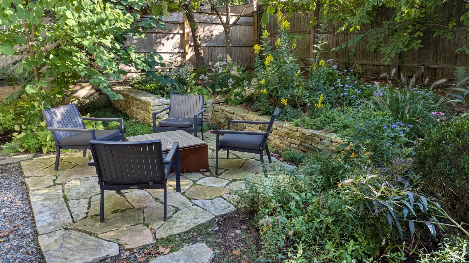 A backyard seating area with four black chairs surrounding a square fire pit, set on a stone patio. The background features a variety of lush green plants, flowers, and trees, with a wooden fence enclosing the garden.