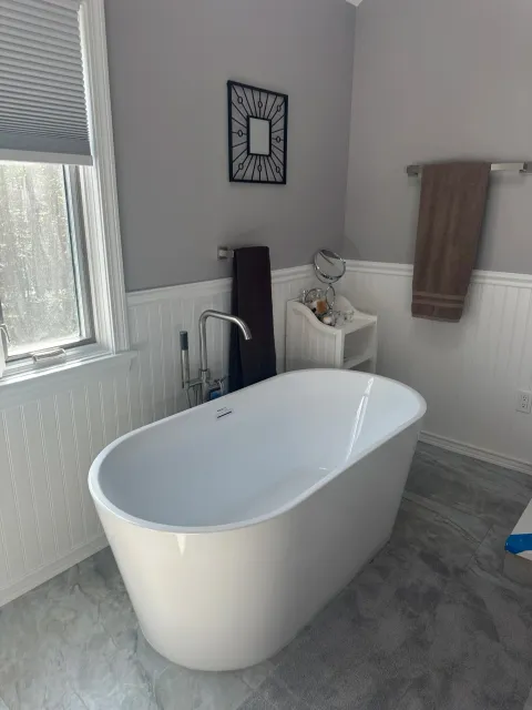 Modern bathroom with freestanding white bathtub, gray floor tiles, gray painted walls, a window with blinds, towel racks with brown towels, and wall art.