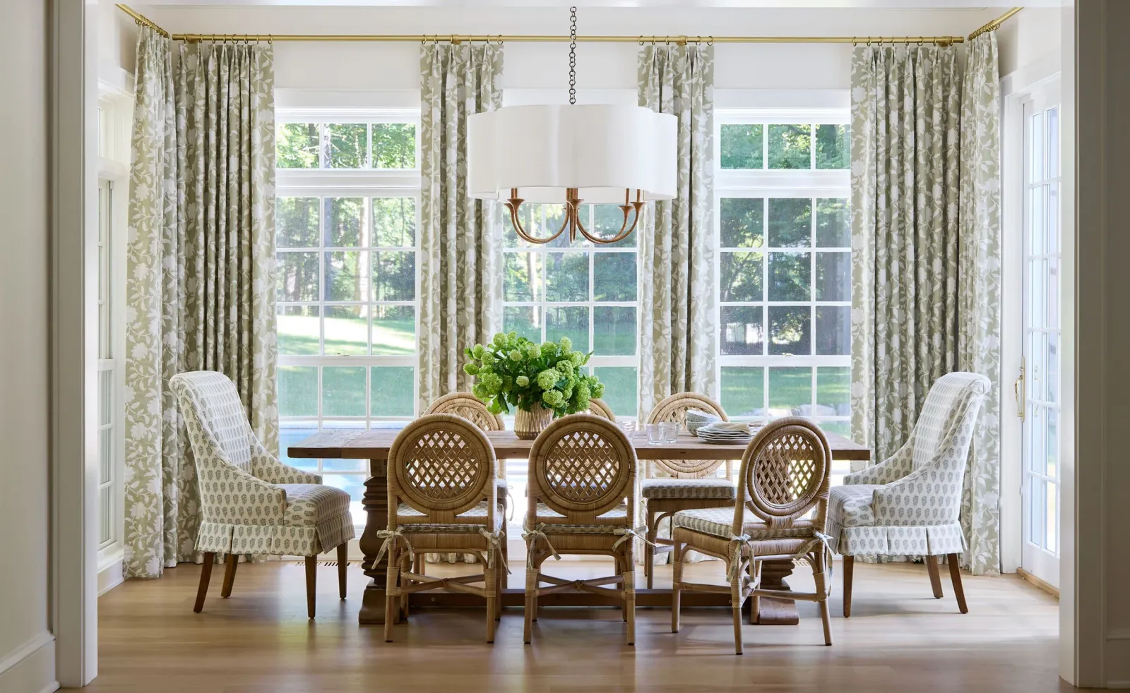 Elegant dining room with wooden table, wicker chairs, patterned curtains, large windows, and a modern chandelier. Timeless, fresh design, and garden inspired