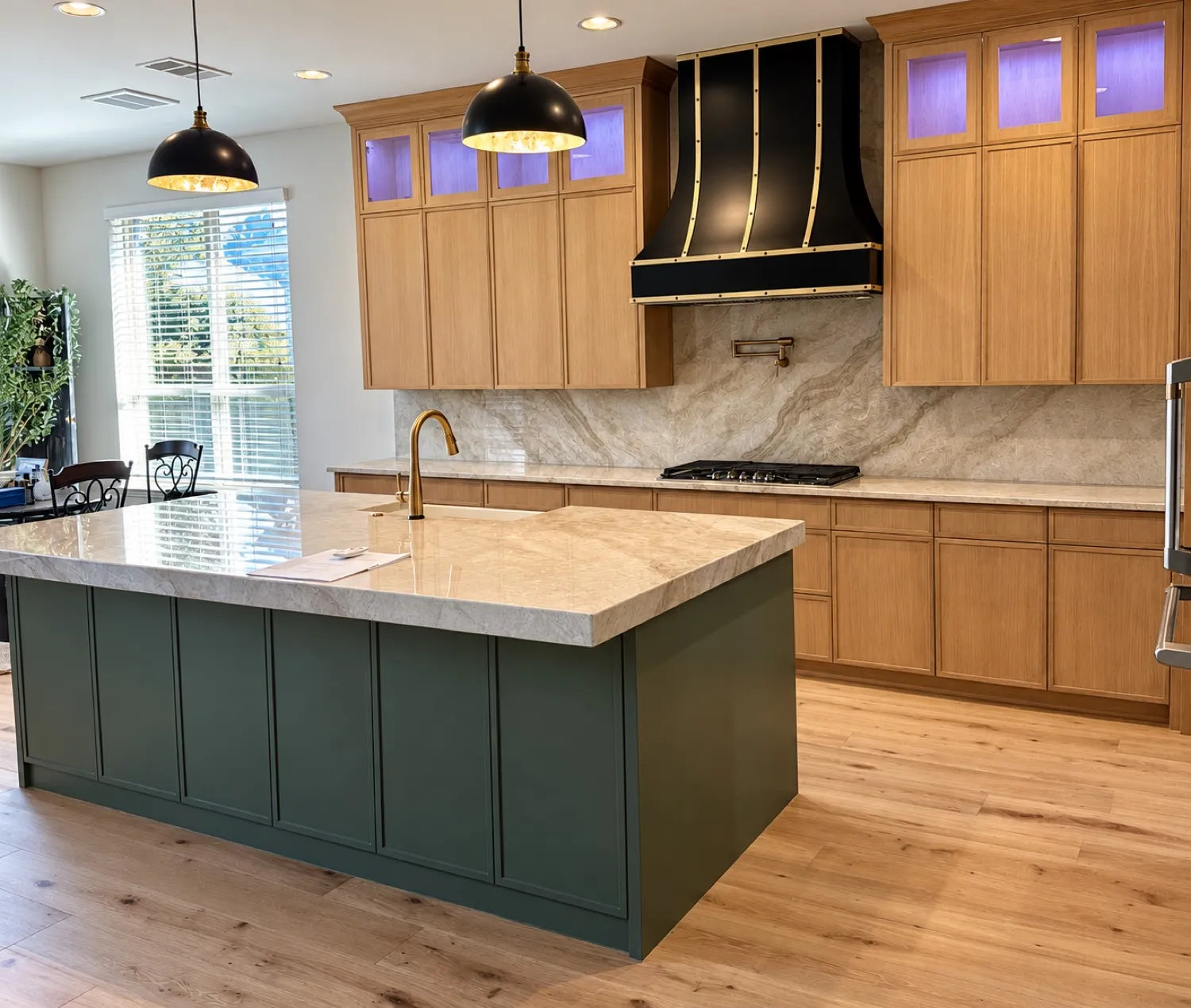 The same kitchen after renovation by Capstone Design Partners: white-oak cabinetry, a deep green island with a quartzite waterfall counter, a black and brass range hood, stone slab backsplash and wide-plank oak floors