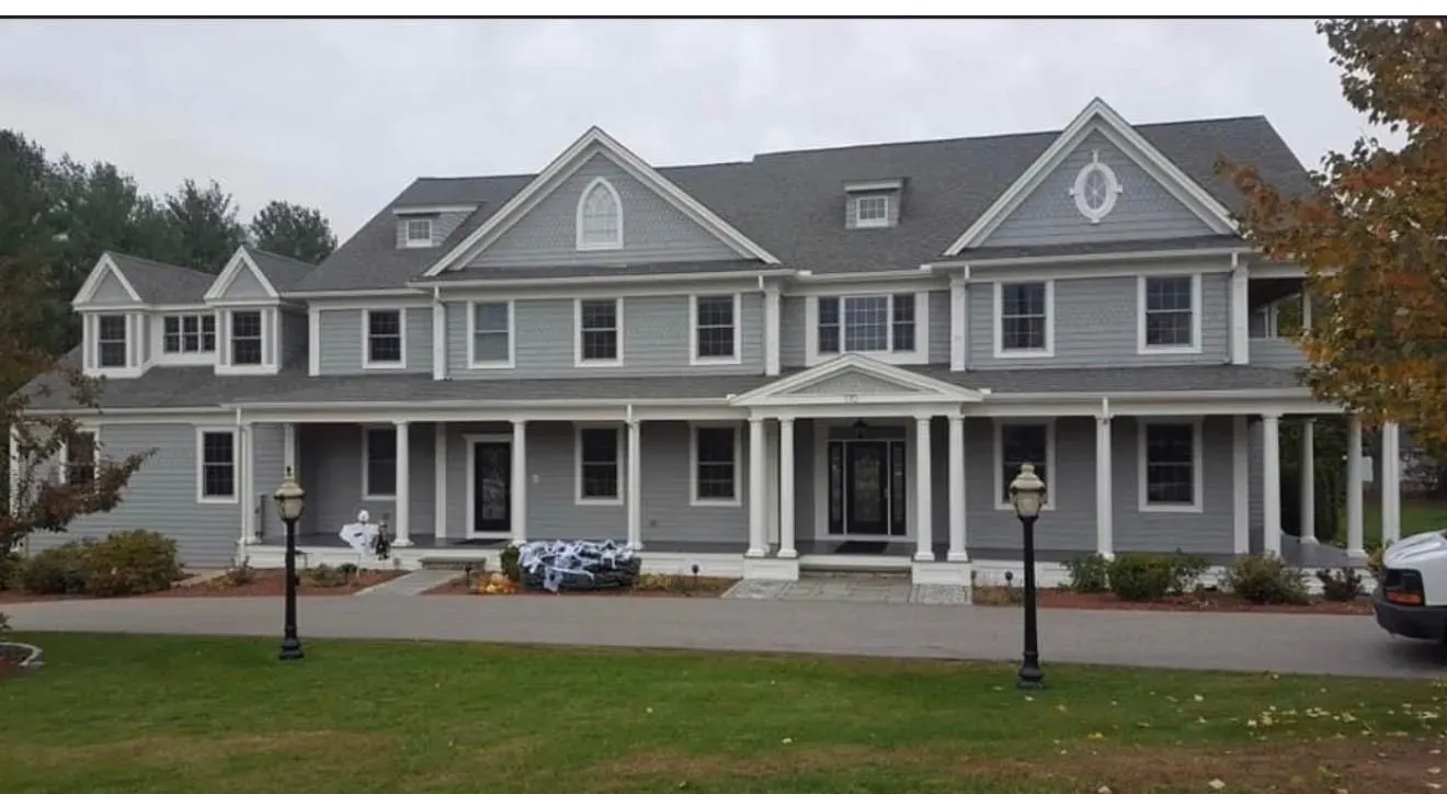 Large gray two-story house with white trim, front porch, dormer windows, and lawn under an overcast sky