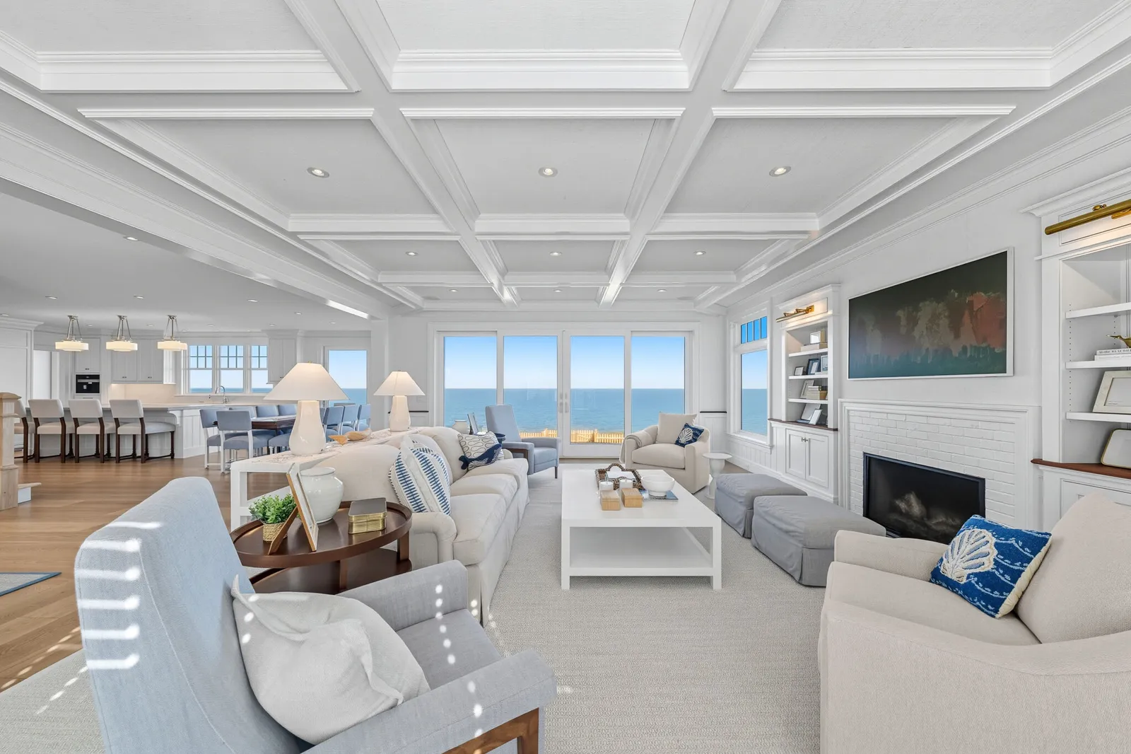 Spacious open-plan living room in a custom Cape Cod home with coffered ceilings, floor-to-ceiling windows with ocean views, white fireplace, built-in shelving, and coastal-style furnishings