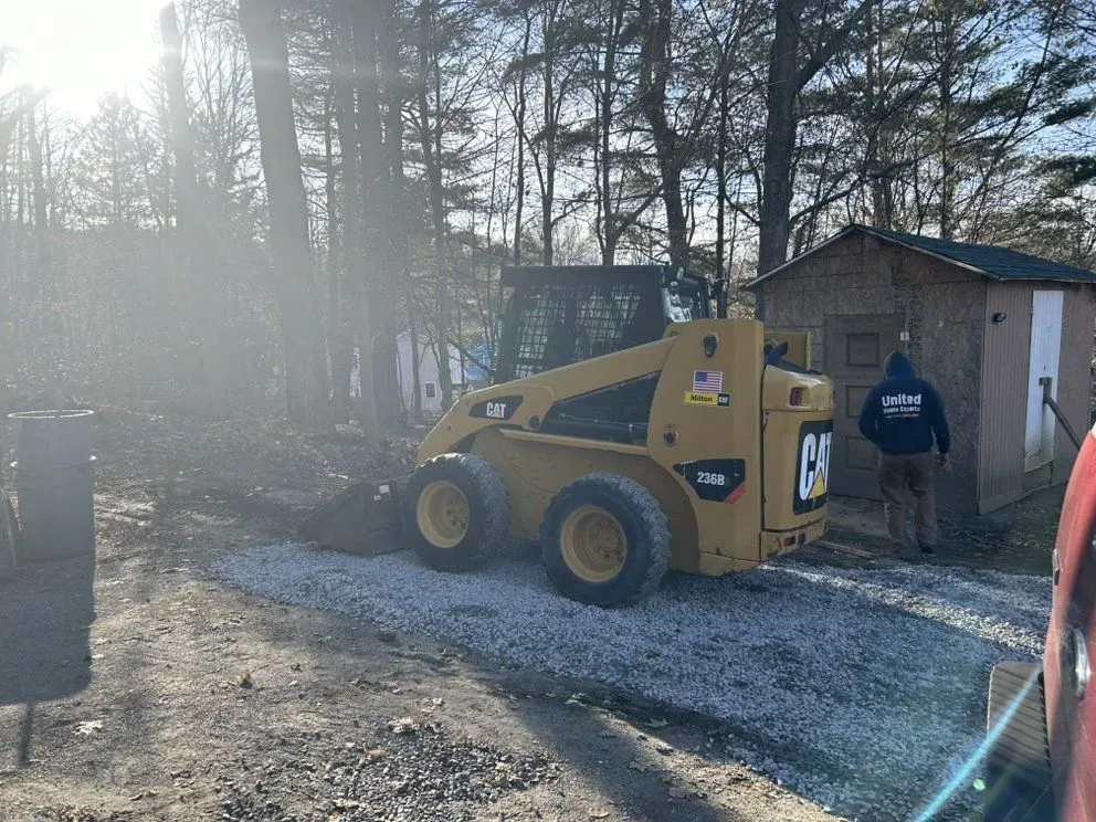 Yellow skid steer beside a shed on a sunny gravel driveway, with two people nearby
