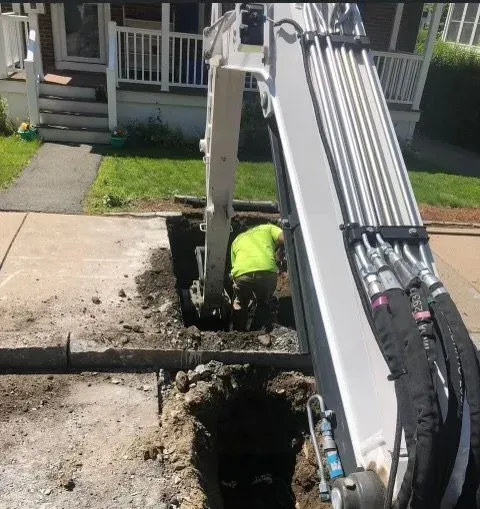 A worker operating a small excavator digging a trench in a residential yard near a sidewalk and a house with a porch.