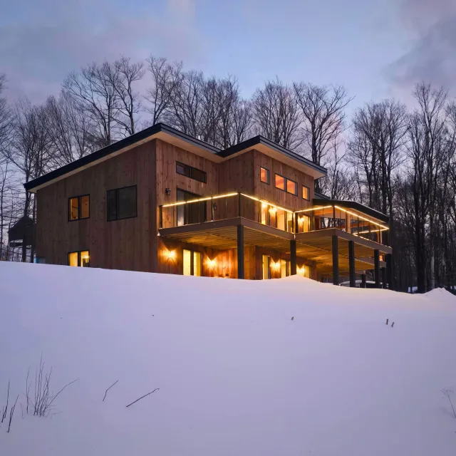 Warm light in the windows of the Killington timber frame home, surrounded by snow and bare winter trees.