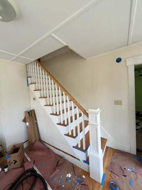 Interior view of a staircase under renovation, with white painted balusters and natural wood handrail, area covered in protective paper and painter's tape, debris and tools scattered on the floor.