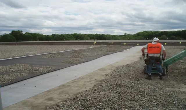 Worker in orange vest operating a machine on a gravel road beside a concrete drainage channel.
