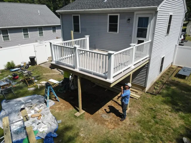 Backyard deck with white railing beside a house; a worker stands below near construction materials.