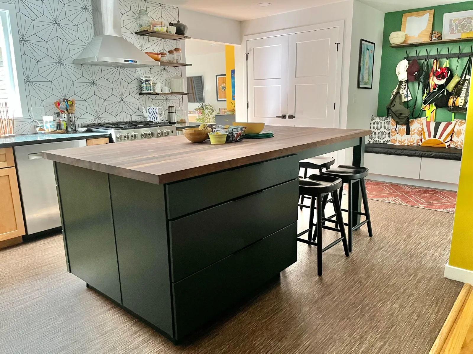 Modern kitchen with a large black island countertop, wooden surface, bar stools, open shelving, patterned wall, and a cozy entryway with hooks, backpacks, and a bench with cushions.