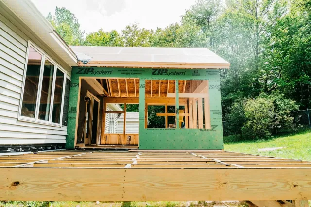 Partially built green house addition beside a white home with exposed framing and wooden deck floor