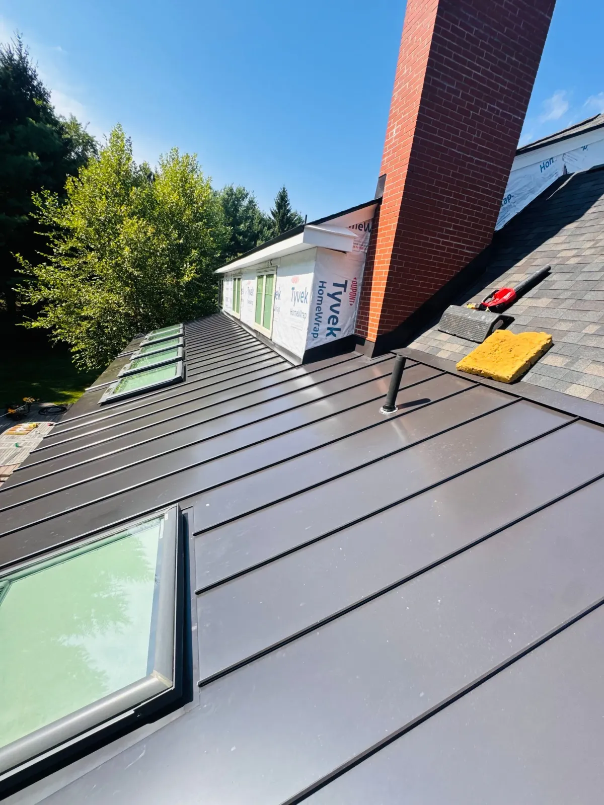 Sloped gray metal roof with skylight, red chimney, and yellow tool near the ridge under a blue sky