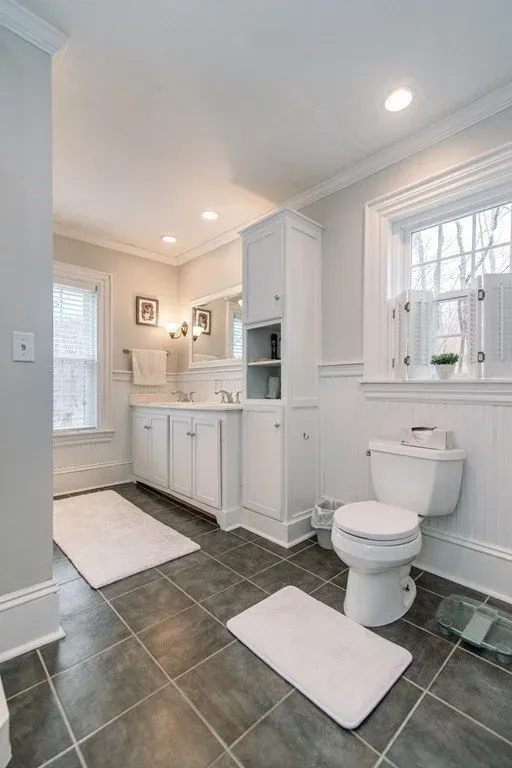 Bright white bathroom with large window, white vanity with under-sink storage, toilet, dark tiled floor, white mats, and small potted plant on windowsill.