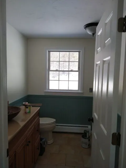 Small bathroom with white walls, green wainscoting, a window, toilet, sink cabinet, and open door.
