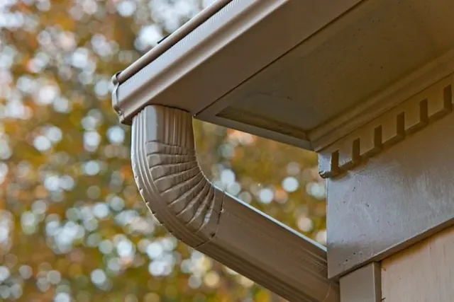 Close-up of a beige gutter and downspout on the side of a house with autumn trees in the background.