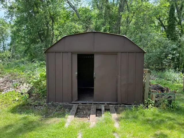 Brown metal shed with an open doorway in a wooded backyard, with a short ramp leading up to it