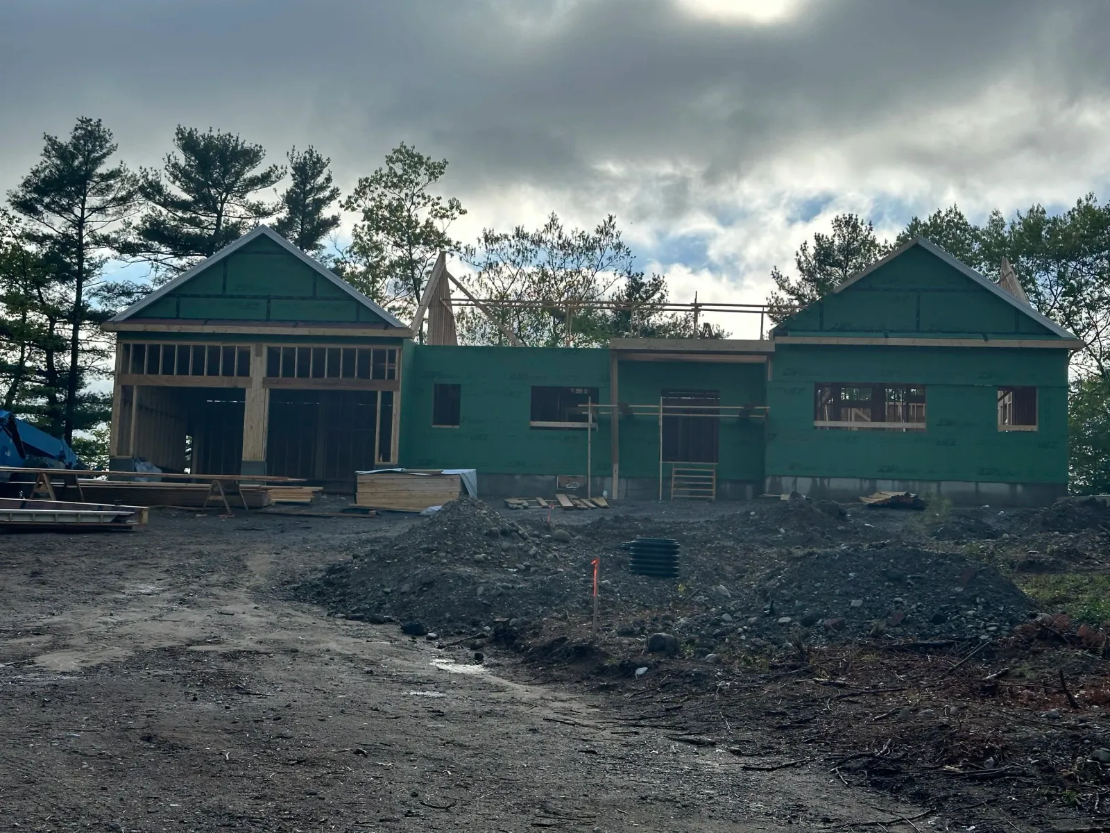 A house under construction with a partially built wooden frame and green siding, surrounded by construction materials and piles of dirt, with a backdrop of trees and a cloudy sky.